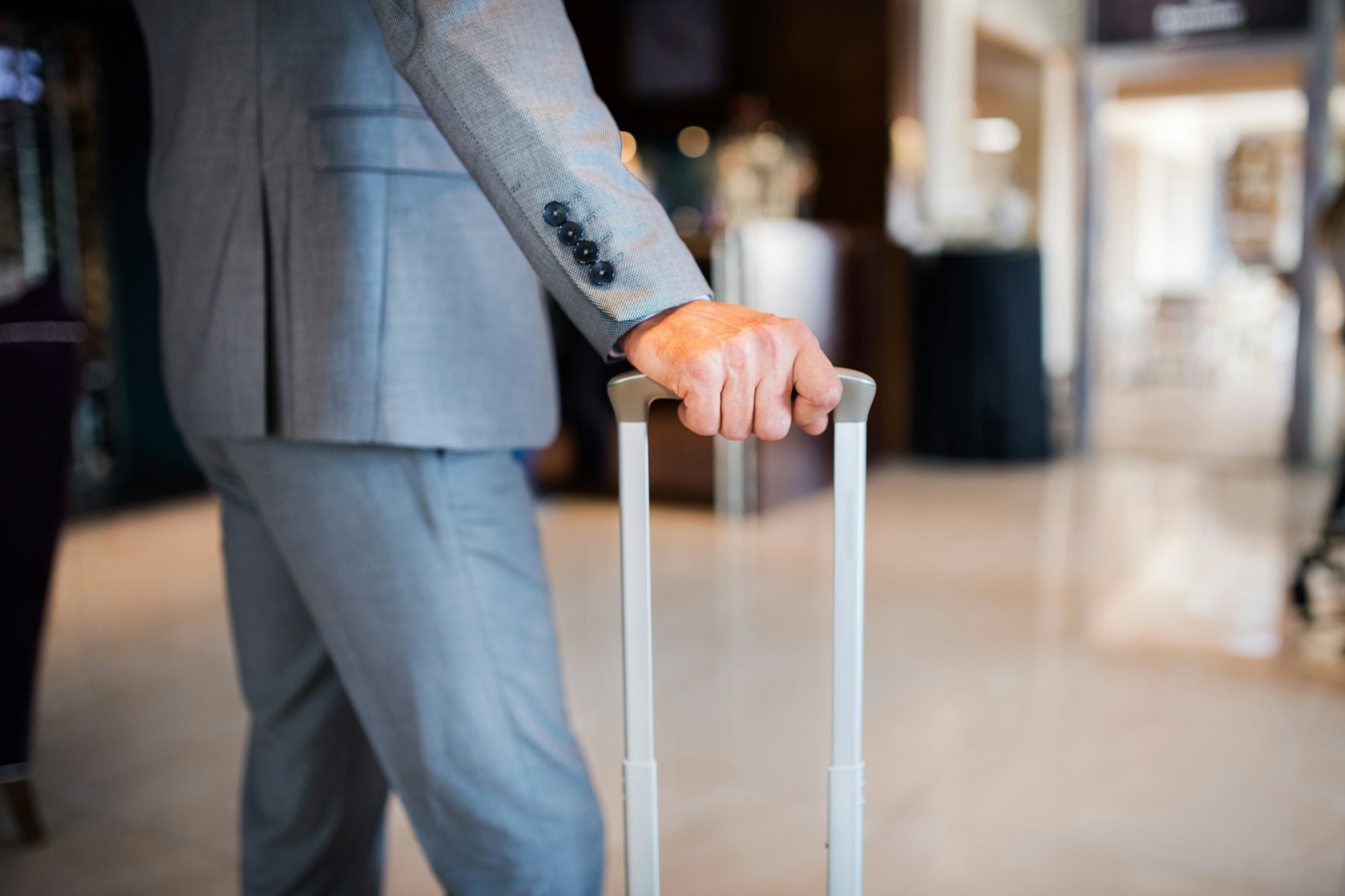 A business traveler in a suit gripping the handle of a rolling suitcase in a hotel lobby